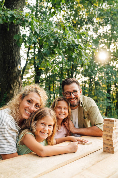 Retrato de uma família feliz de quatro pessoas sentadas na natureza à mesa e abraçando enquanto sorri para a câmera. Jovens pais carinhosos abraçando suas filhas e fazendo contato visual. Espaço de cópia. - Foto, Imagem