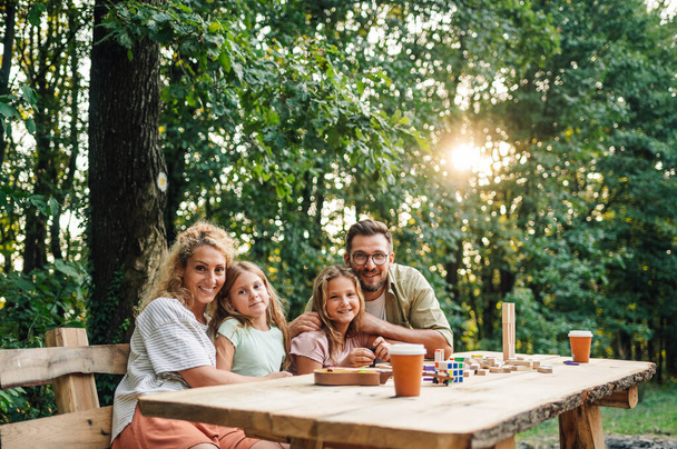 Eine liebevolle vierköpfige Familie sitzt am Picknicktisch in der Natur und umarmt sich, während sie in die Kamera lächelt. Junge Eltern umarmen ihre Töchter und sitzen am Picknicktisch in der Natur. Kopierraum. - Foto, Bild