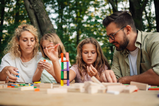 Birlikte doğada piknik masasında tahta blok oyunu oynayan eğlenceli bir ailenin portresi. Doğada birlikte oyun oynayan dört kişilik bir aile. Aile bloklardan bir kule inşa ediyor. - Fotoğraf, Görsel