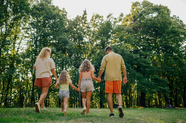 Back view of an adventurous family of four holding hands and exploring nature together. A young explorer family is hiking in nature. Rear view of family of four holding hands and exploring nature. - Photo, Image