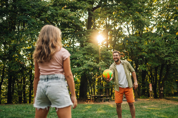 Een sportieve vader serveert een bal en speelt volleybal in de natuur met zijn dochter. Pap en dochter die op zonnige zomerdag in de natuur sporten. Sportieve familie met een bal. - Foto, afbeelding