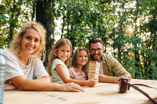 ピクニックのテーブルに座って,自然の中で楽しいジェンガボードゲームをプレイする家族. カメラで笑顔ながら楽しい木のブロックパズルゲームをプレイする自然の中で陽気な家族の日を過ごす肖像画. - 写真・画像