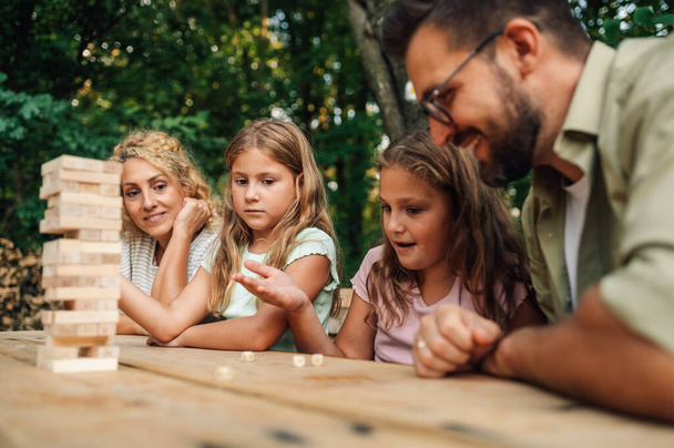 A happy family is sitting at the table in nature on picnic and playing fun jenga board game. A girl is rolling dices and planing her next move while playing a wood block puzzle game with her family. - Photo, Image