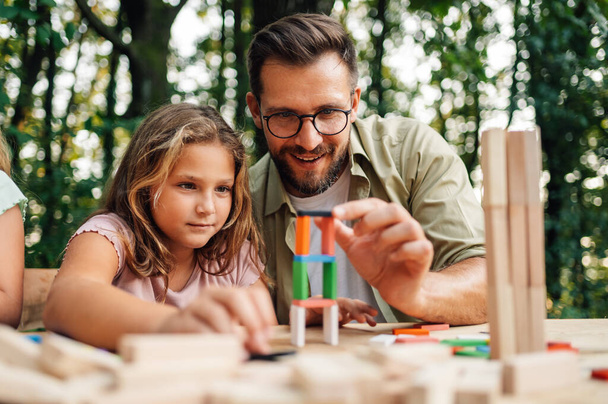 自然の中で娘と楽しい教育ゲームをしている父親の肖像画. 家族はピクニックのテーブルに座り,カラフルな木製タイルの固体構造を構築しています. ボードゲームデー. - 写真・画像