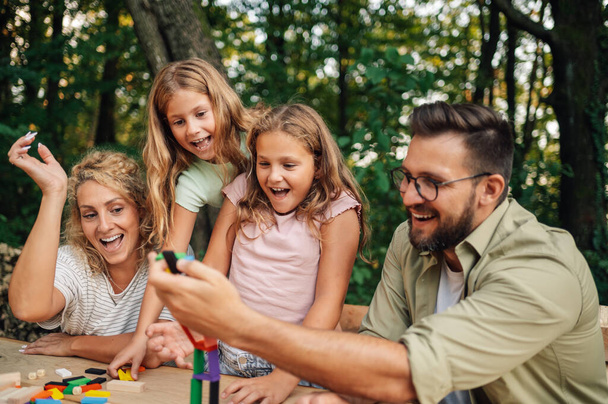A playful happy family is sitting at the table in nature and playing a fun board game. A wood tiles structure is crumbling while family is laughing and having a great time together. Fun playtime. - Photo, Image
