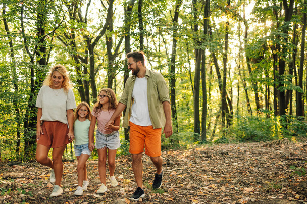 Szczęśliwi rodzice trzymają się za ręce ze swoimi córkami i spacerują po lesie. Aktywna kochająca się rodzina spędza razem dzień w przyrodzie. Portret aktywnej rodziny spacerującej w lesie w słoneczny dzień. - Zdjęcie, obraz