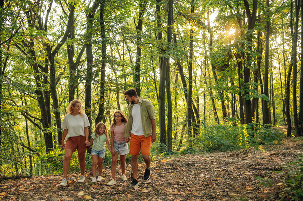 Wesoła rodzina trzyma się za ręce i bada las. Portret odważnych rodziców z córkami spacerujących po przyrodzie i spędzających dzień w lesie w słoneczny dzień. Rodzina w lesie. Przestrzeń kopiowania. - Zdjęcie, obraz