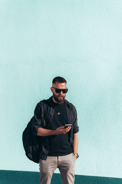 Joven hombre guapo usando un teléfono inteligente y usando gafas de sol afuera con una pared blanca en el fondo - Foto, Imagen