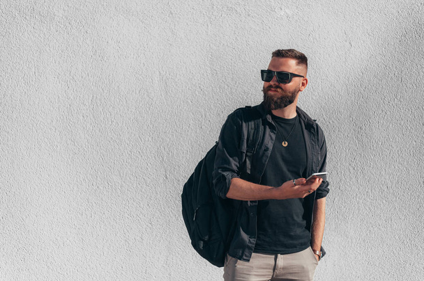Akıllı telefon kullanan, sıradan giyinen ve arka planda beyaz bir duvar olan güneş gözlüğü takan genç yakışıklı bir adam. - Fotoğraf, Görsel