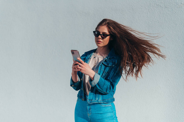 Jonge mooie vrouw met behulp van een smartphone en het dragen van jeans en een zonnebril terwijl ze voor een witte muur - Foto, afbeelding