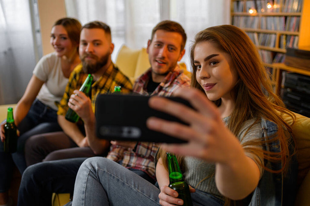 Skupina přátel, kteří se spolu poflakují na večírku, berou selfie s chytrým telefonem a pijí pivo, zatímco se baví - Fotografie, Obrázek