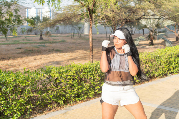 Donna con un top trasparente e cappuccio bianco assume una posizione di pugilato in un parco soleggiato, concentrandosi sulla sua sessione di allenamento. - Foto, immagini
