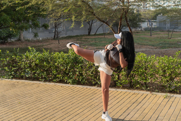 Vrouw met een transparante boven- en witte pet die een hoge kick geeft tijdens een outdoor bokssessie in een zonnig park, met flexibiliteit en kracht. - Foto, afbeelding
