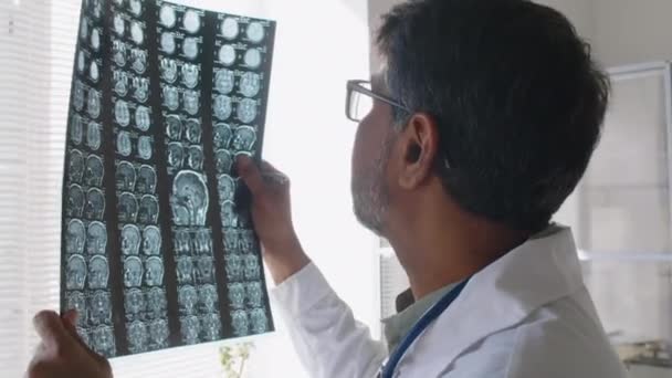 Gros plan d'un oncologue asiatique d'âge mûr, concentré et fronçant les sourcils, étudiant les résultats de la tomographie par IRM cérébrale sur film par fenêtre en clinique, détectant la tumeur et la traçant avec un stylo - Séquence, vidéo