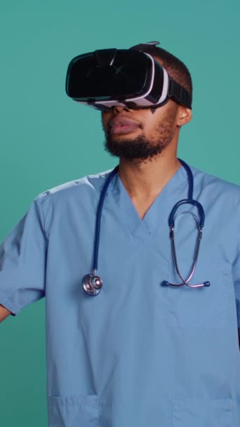 Вертикальное видео медсестра носит наушники виртуальной реальности удаленно оперирующий пациент. Специалист по здравоохранению с использованием высокотехнологичного современного медицинского гаджета vr, изолированный на фоне синей студии, камера - Кадры, видео