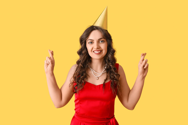 jonge vrouw in verjaardag hoed maken wens op gele achtergrond - Foto, afbeelding