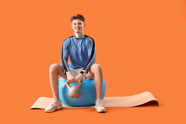 Sportowy młody człowiek z kettlebell na fitball na pomarańczowym tle - Zdjęcie, obraz