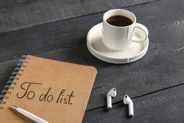 Notebook with text TO DO LIST, cup of coffee and earphones on wooden black background - Photo, Image
