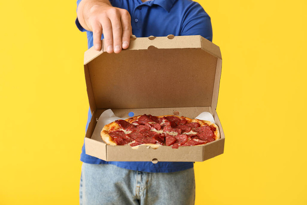 Young man holding cardboard box with tasty pepperoni pizza on yellow background, closeup - Photo, Image