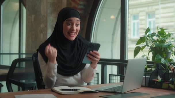 Αραβικός ισλαμικός μουσουλμάνος γυναίκα των επιχειρήσεων σε hijab κατάπληκτος χαρούμενος κορίτσι γυναίκα ενθουσιασμένος επιχειρηματίας κινητό τηλέφωνο καλά νέα κερδίσει smartphone στοίχημα σε απευθείας σύνδεση γιορτάσουμε κραυγή φωνάξει ναι επιτυχία επίτευγμα στο γραφείο - Πλάνα, βίντεο
