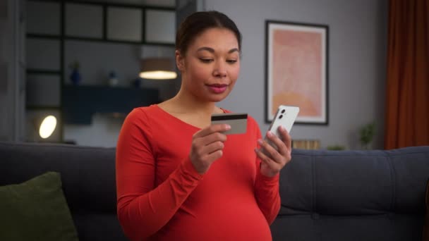 Happy smiling pregnant African American woman girl female mother at home couch buying order food delivery cloth store shop online internet payment pay mobile phone credit card bank purchase shopping - Footage, Video