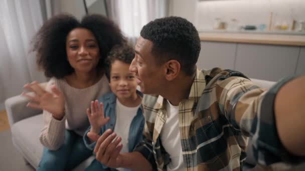 Boldog család afro-amerikai férfi nő férfi nő szülők kisfiú gyerek fia néz kamera online telefon okostelefon mobil videó hívás chat app beszél integető kéz gesztus köszöntés beltéri szobában - Felvétel, videó