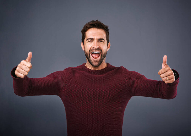 Man, success and thumbs up in studio for winning, support and like or yes for achievement. Portrait of excited person shouting, cheering and motivation with good luck and voting on gray background. - Photo, Image