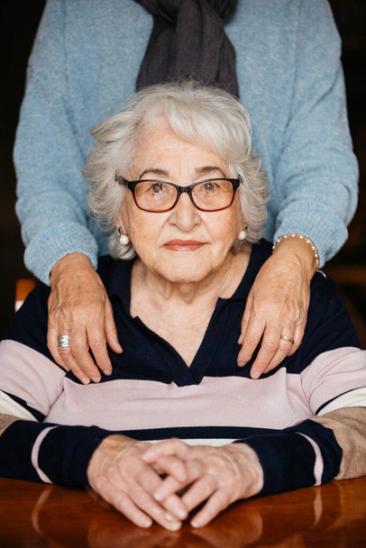Προσωπογραφία ηλικιωμένης γυναίκας που κάθεται στο τραπέζι με τα χέρια σταυρωμένα, ενώ τα χέρια της κόρης της αναπαύονται απαλά στους ώμους της, δείχνοντας συμπαράσταση και στοργή. - Φωτογραφία, εικόνα
