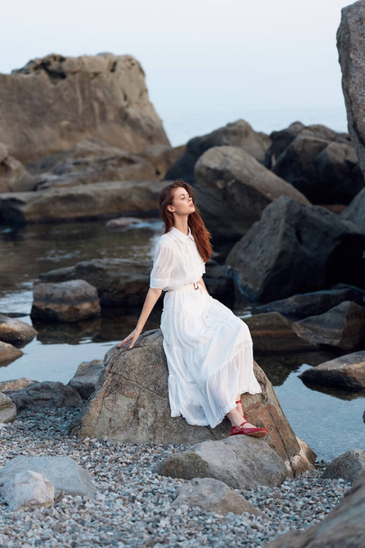 Смертельна жінка в білій сукні, сидячи на скелястому узбережжі океану з руками, витягнутими у мирному спогляданні - Фото, зображення