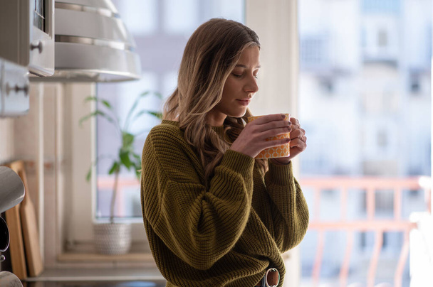 Çekici genç bir kadın mutfakta duruyor ve bir fincan çay içiyor. Rahatlama anı. Mutfakta sabah kahvesinin tadını çıkarıyor.. - Fotoğraf, Görsel