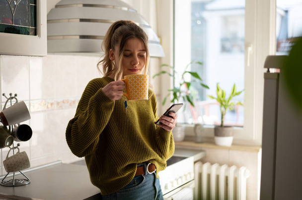 Красива жінка, використовуючи смартфон на кухні, п'є чашку свіжого чаю. Молода жінка здоровий спосіб життя вдома вранці. - Фото, зображення
