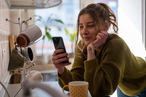 Mujer joven atractiva usando un teléfono inteligente y beber una taza de té mientras está de pie en la cocina en casa. Concepto de actividad de ocio en el hogar. - Foto, imagen
