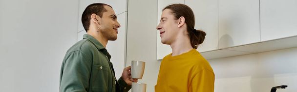 Mladý gay pár si užívá neformální dopoledne ve své moderní kuchyni, sdílí teplý drink a usmívá se. - Fotografie, Obrázek