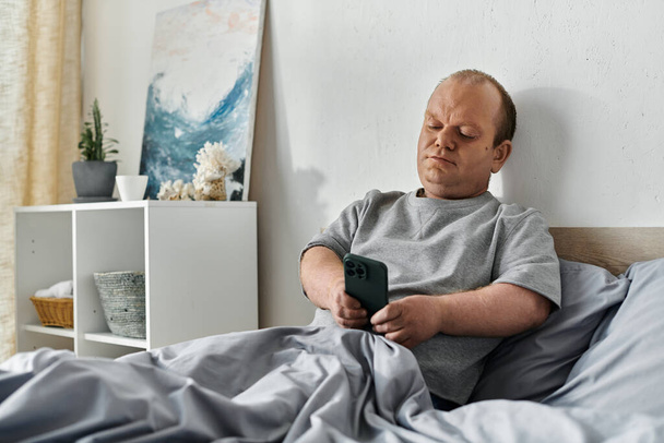 Muž s inkluzivitou sedí v posteli a dívá se na svůj telefon.. - Fotografie, Obrázek