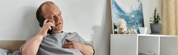Un homme avec inclusivité est assis sur un lit, parlant au téléphone, dans une pièce lumineuse et aérée. - Photo, image