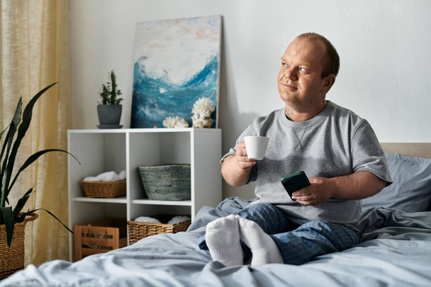 Mężczyzna z inkluzywnością siedzi w łóżku, pijąc kawę i patrząc przez okno, może zastanawiając się nad swoim dniem.. - Zdjęcie, obraz