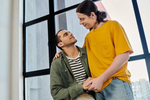 Ένα νεαρό γκέι ζευγάρι μοιράζεται μια τρυφερή στιγμή στο μοντέρνο διαμέρισμά του, γεμάτο φυσικό φως.. - Φωτογραφία, εικόνα