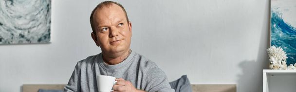 Um homem com inclusividade em uma camisola cinza senta-se em um sofá, segurando uma caneca de café. - Foto, Imagem