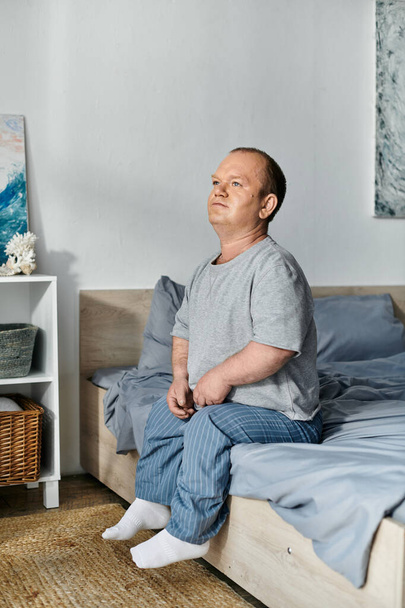 Egy inkluzivitással teli férfi ül az ágya szélén, elveszve a gondolataiban, ahogy a reggeli fény beáramlik. - Fotó, kép