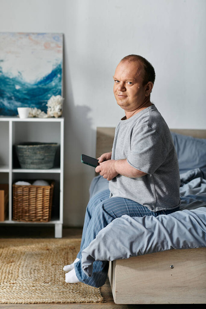 Un hombre con inclusividad se sienta en su cama, mirando su teléfono. Lleva ropa casual y parece relajado.. - Foto, Imagen