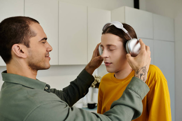 A young gay couple relaxes in their modern apartment, one putting headphones on the other. - Photo, Image