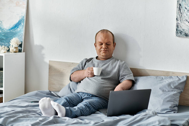 Un hombre con inclusividad se sienta en la cama, disfrutando de una taza de café mientras usa su computadora portátil. - Foto, imagen