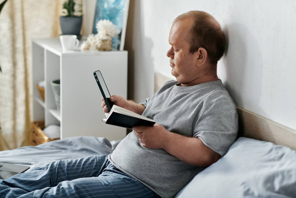 Een man met inclusiviteit zit in bed en kijkt naar zijn telefoon terwijl hij een boek vasthoudt.. - Foto, afbeelding