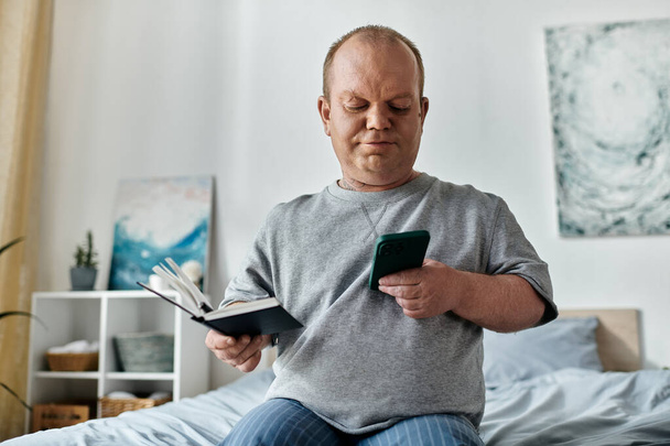 Un homme avec inclusivité est assis sur un lit, tenant un livre et vérifiant son téléphone. - Photo, image