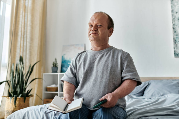 Een man met inclusiviteit zit op een bed, houdt een boek en een telefoon vast, kijkt bedachtzaam vooruit. - Foto, afbeelding