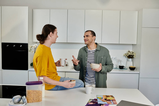 Ένα νεαρό γκέι ζευγάρι απολαμβάνει ένα χαλαρό πρωινό στο μοντέρνο διαμέρισμά του, πίνοντας ένα φλιτζάνι καφέ και ένα μπολ δημητριακά.. - Φωτογραφία, εικόνα