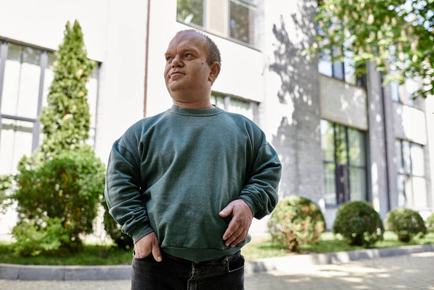 Ένας άντρας με εγκλειστικότητα στέκεται με αυτοπεποίθηση έξω από ένα κτίριο, περιτριγυρισμένος από πράσινο, που ενσωματώνει το πνεύμα της εγκλειστικότητας.. - Φωτογραφία, εικόνα