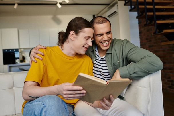 Młoda para gejów dzieli się śmiechem czytając razem książkę w nowoczesnym mieszkaniu. - Zdjęcie, obraz