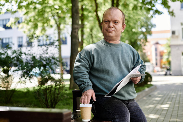 Un homme inclusif s'assoit sur un banc dans un parc, lit un journal et prend une tasse de café. - Photo, image