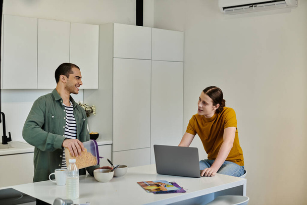 Una joven pareja gay disfruta de una mañana informal en su apartamento moderno, con uno preparando el desayuno, mientras que el otro trabaja en un ordenador portátil. - Foto, imagen